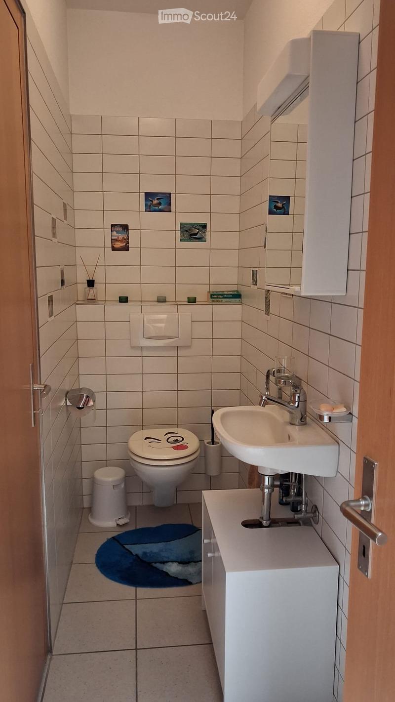 Compact bathroom with white tiled walls and floor, featuring a toilet with a smiley face design, a sink with a modern faucet, a mirror cabinet, and a small trash bin.