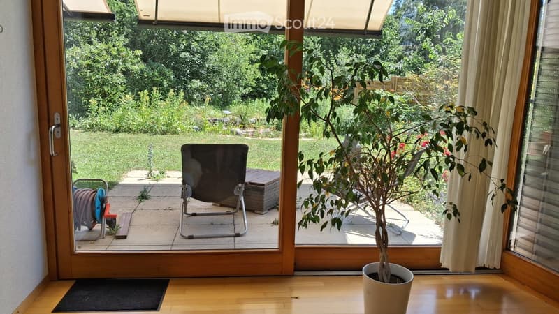 A spacious balcony with a wooden floor, a potted plant, a folding chair, and a small table, overlooking a lush green garden.