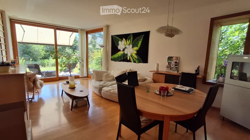 Bright living and dining area with wooden flooring, large windows, a sofa, coffee table, dining table with chairs, and a lamp.