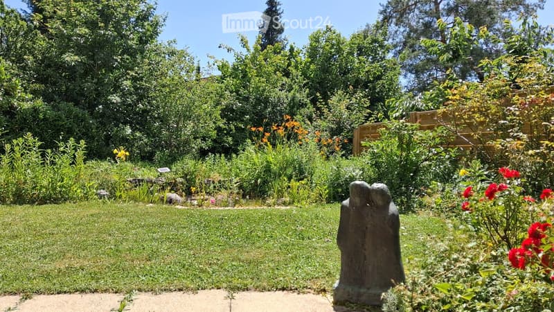 Well-maintained garden with a variety of plants, flowers, and a sculpture of two figures. The garden features a neatly trimmed lawn, colorful blooms, and a wooden fence in the background.