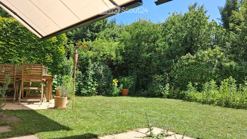 A well-maintained garden with a wooden table and chairs, surrounded by lush greenery and potted plants, under a covered patio area.