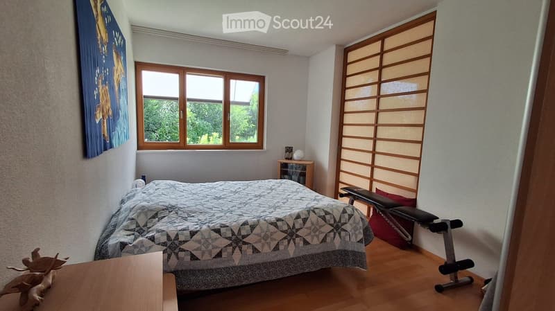 Cozy bedroom with wooden flooring, large window, bed with patterned quilt, nightstand, exercise bike, small table with a decorative statue, and a colorful poster on the wall.
