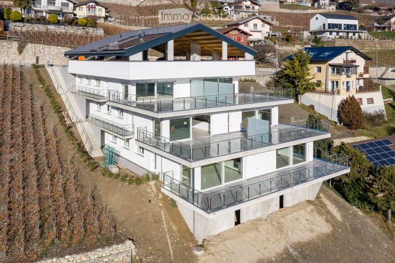 Modern two-story house with large glass balconies and a flat roof, situated on a hillside with a scenic view of the surrounding area.