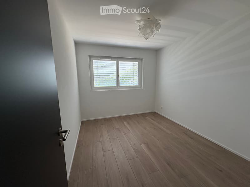 Empty room with wooden flooring, white walls, and a single window with blinds. The room features a modern light fixture on the ceiling and a door on the left side.