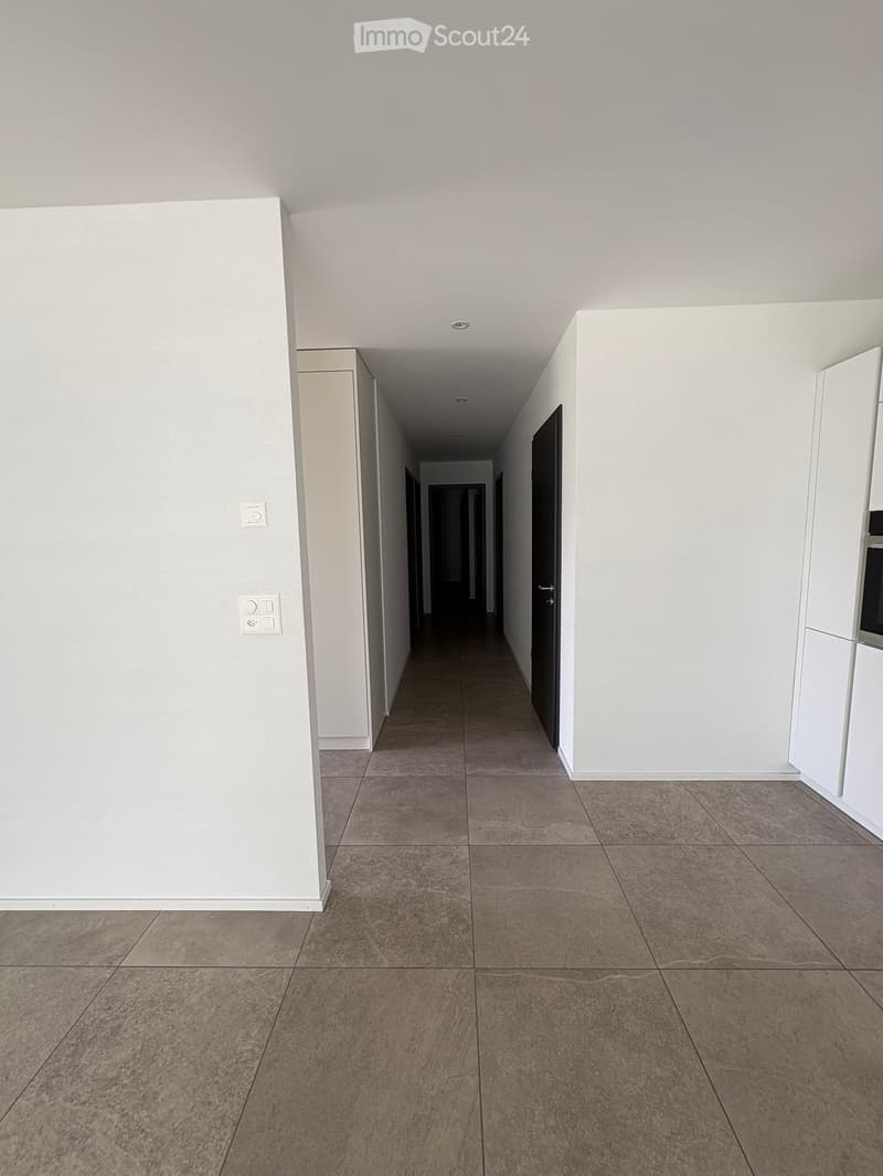 Empty hallway with tiled floor, white walls, and recessed lighting. Doors visible at both ends.