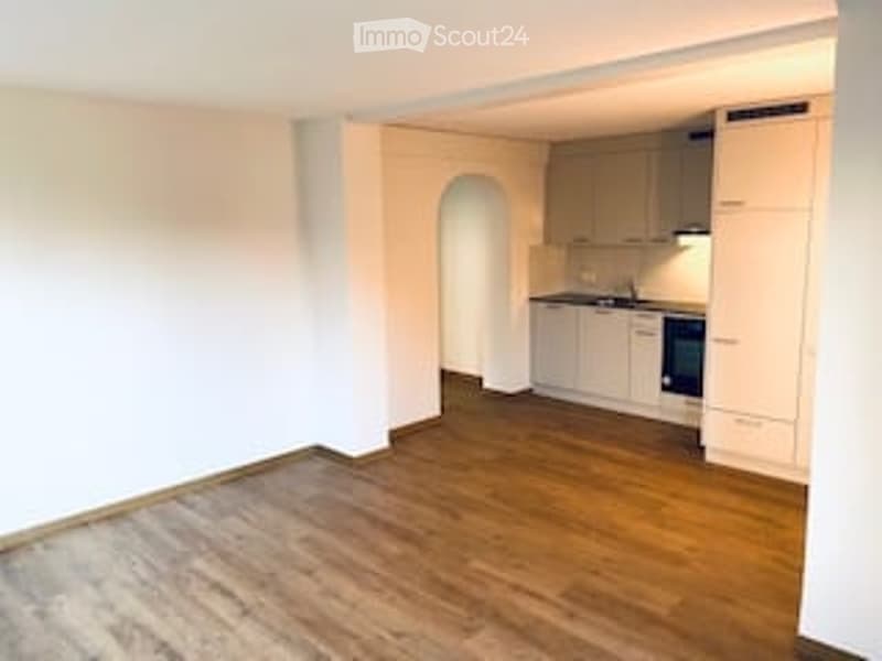 Empty room with wooden flooring, white walls, and a small kitchen area with cabinets and appliances visible in the background.