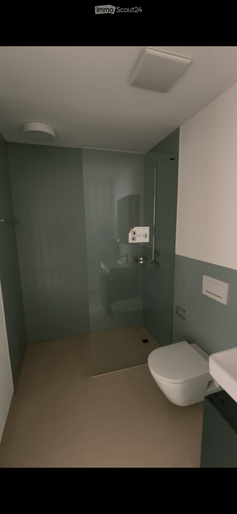 Modern bathroom with a toilet, sink, and glass-enclosed shower. Dark green tiles on walls, light brown tiled floor, and white fixtures.