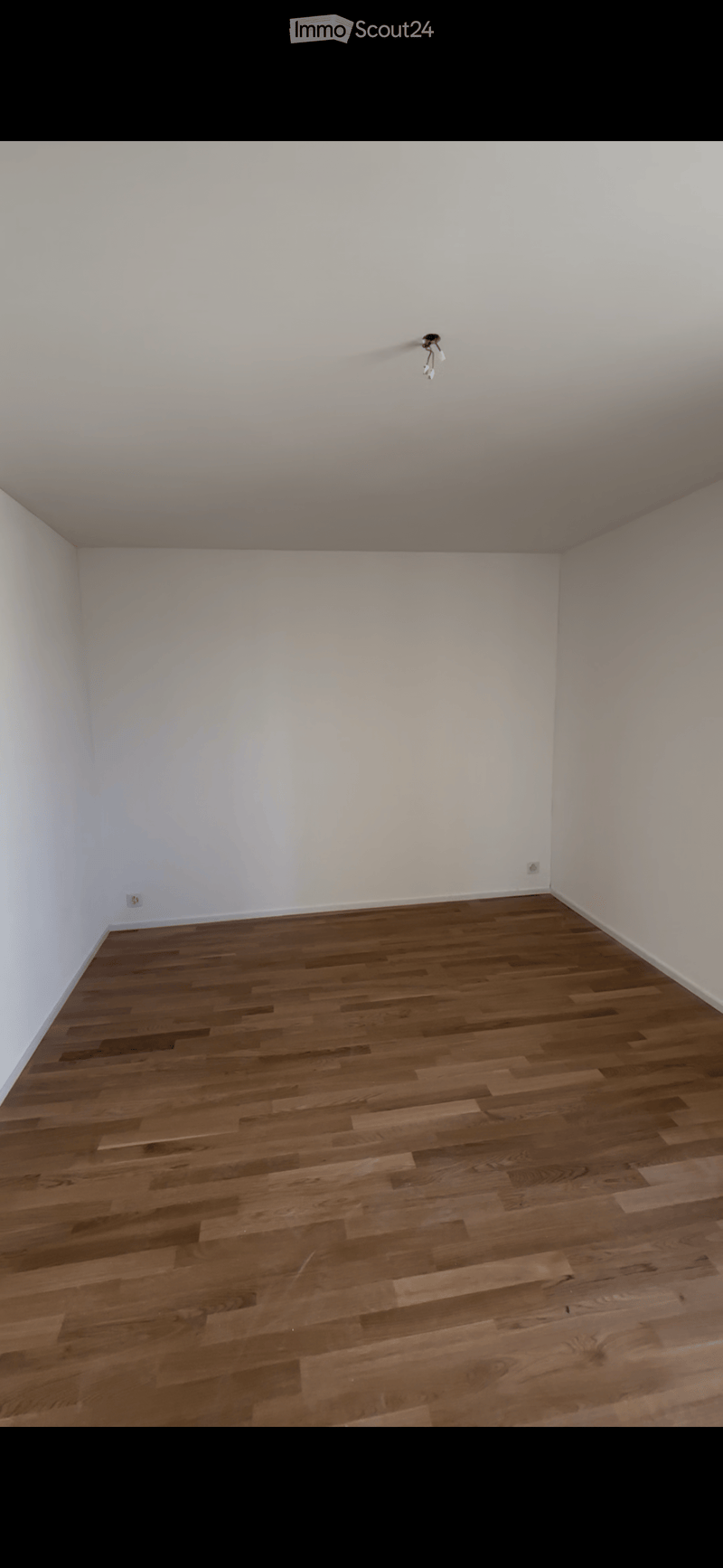 Empty room with white walls and wooden flooring, featuring a single ceiling light fixture and electrical outlets.