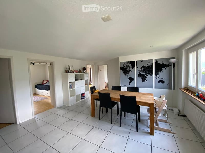 Bright dining area with a wooden dining table, black chairs, a baby chair, a white shelving unit, and a small bedroom visible in the background.