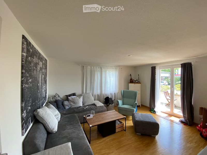Modern living room with grey sectional sofa, wooden coffee table, armchair, ottoman, and large window with curtains. Features a cityscape mural on one wall and a small table with a guitar.