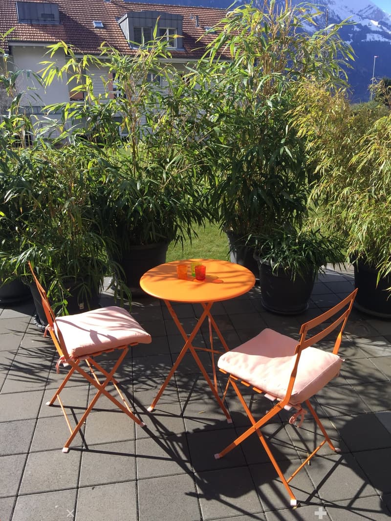 Balcony with a small round table, two chairs, and two cups, surrounded by potted plants and a view of a building and mountains.