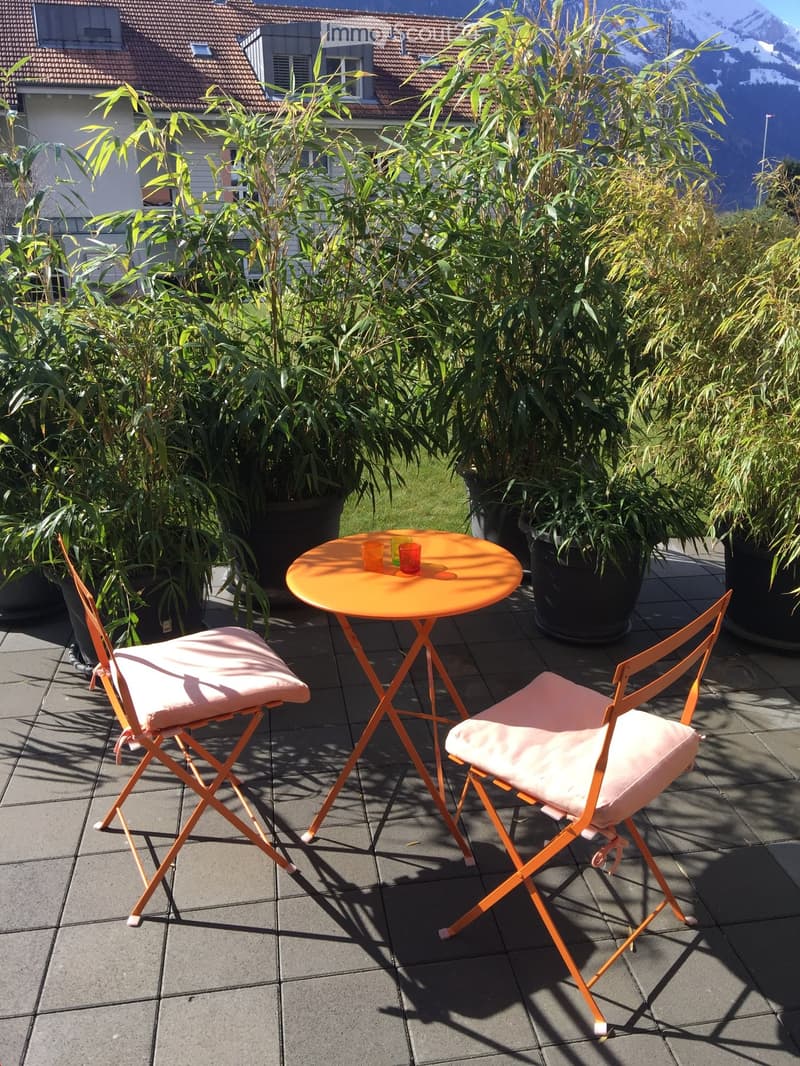 Balcony with a small round table, two chairs, and two cups, surrounded by potted plants and a view of a building and mountains.