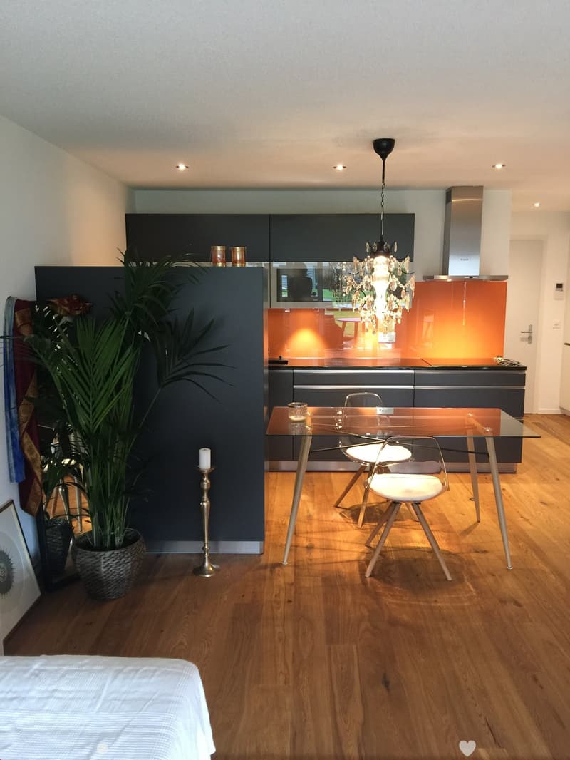 Modern kitchen and dining area with dark grey cabinets, a glass dining table, white chairs, a chandelier, and a potted plant. The floor is wooden, and the kitchen features a stainless steel range hood and a backsplash with an orange accent.