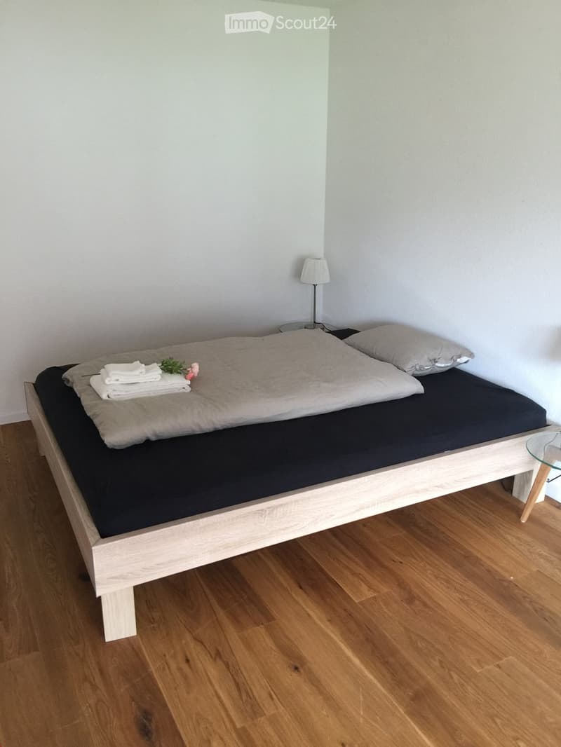A small, neatly arranged bedroom with a single wooden bed, a nightstand with a table lamp, and a small decorative flower arrangement on the bed.