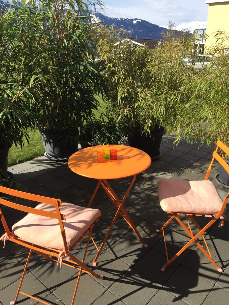 Balcony with a small orange table and two matching chairs, surrounded by potted plants and a scenic mountain view.
