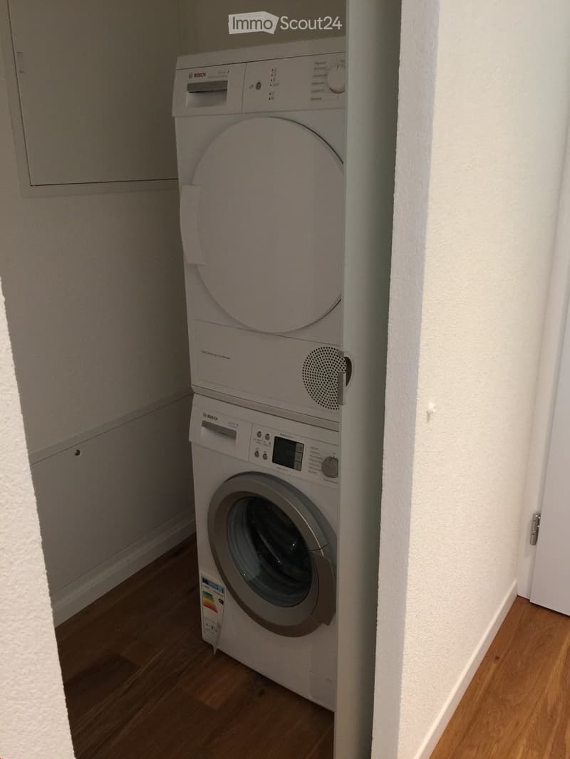 A compact bathroom with a stacked washing machine and dryer unit, white walls, and wooden flooring.