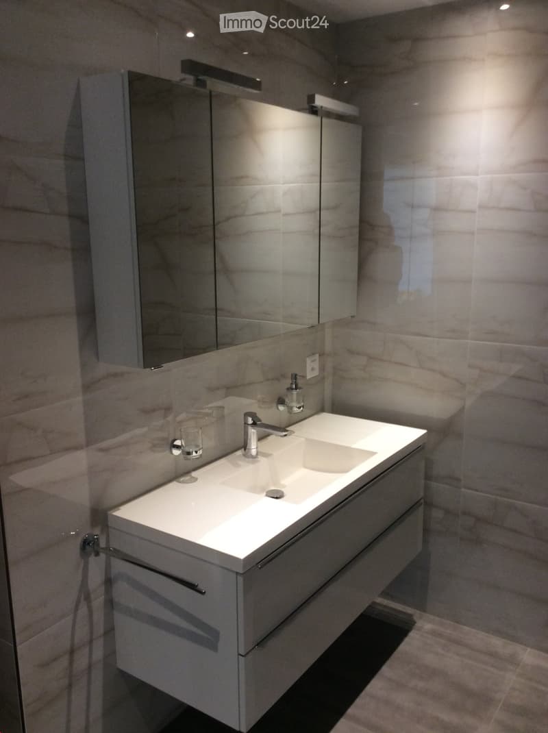 Modern bathroom with a double sink vanity, sleek faucet, large mirror, and built-in storage. The walls are covered in marble tiles, and the floor is tiled.