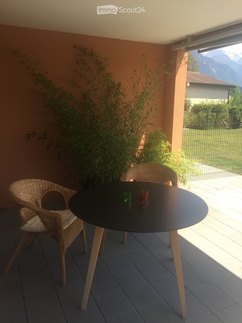 Balcony with a round black table, two wicker chairs, and a scenic mountain view in the background.