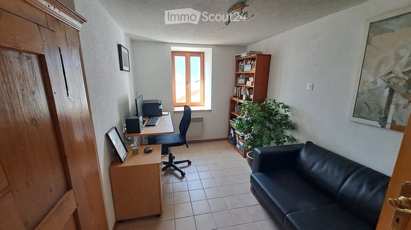 A multi-functional room with a black leather sofa, a wooden desk with a computer setup, a bookshelf, and a potted plant. The room features tiled flooring and a window with wooden shutters.