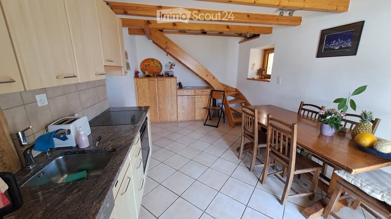 The image shows a cozy kitchen and dining area with a wooden staircase leading to an upper level. The kitchen features a sink, faucet, toaster, and oven, while the dining area includes a wooden table with chairs and a bowl of fruit on the table. A framed
