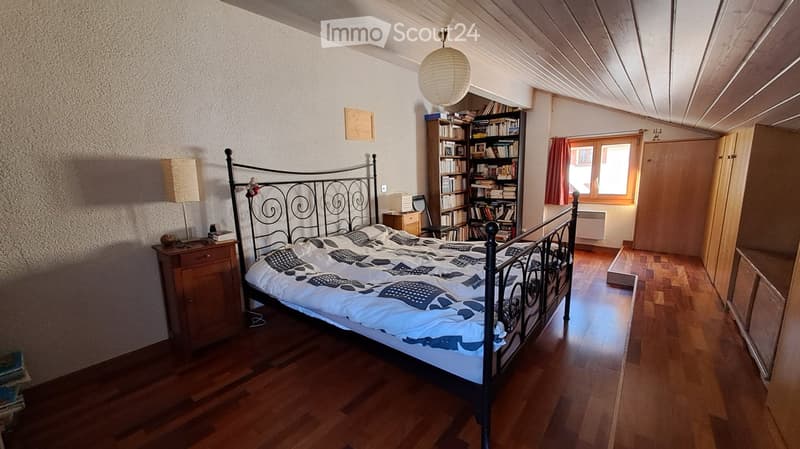 Cozy bedroom with wooden flooring, a metal-framed bed, nightstand, bookshelf, and a window with red curtains.