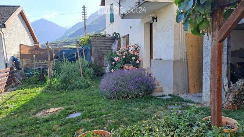 Garden with grass, lavender bushes, rose bushes, potted plants, wooden fence, and a view of mountains in the background.