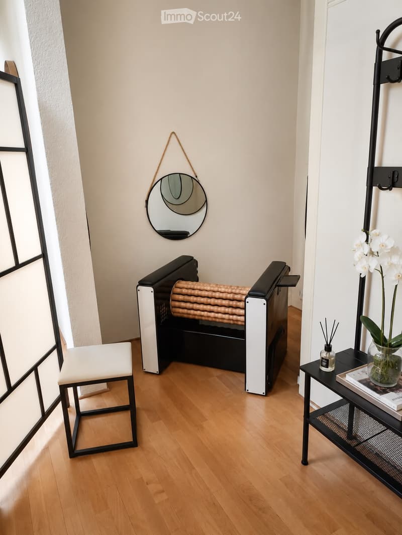 A modern hallway with a wooden floor, a white stool, a black bench, a round mirror, a small black table with a vase of white flowers, and a bottle of perfume.