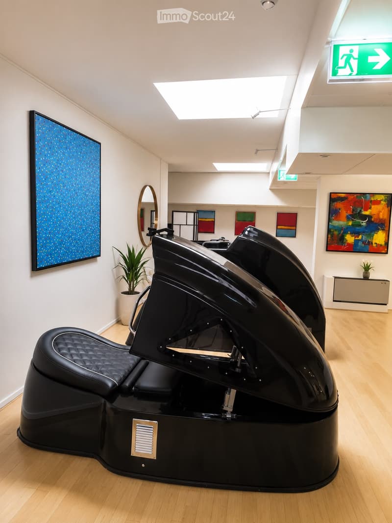 Modern, minimalist room with a black, curved chair, a round mirror, a potted plant, and several pieces of colorful artwork on the walls.
