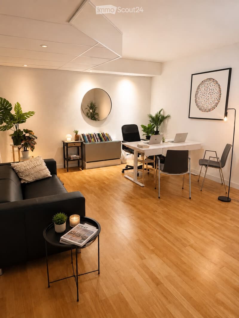Modern, well-lit room with a mix of living and working spaces. Features a black sofa with a white throw pillow, a round coffee table with a candle and magazine, a sleek white desk with two laptops, and several chairs. The room includes a bookshelf, a larg