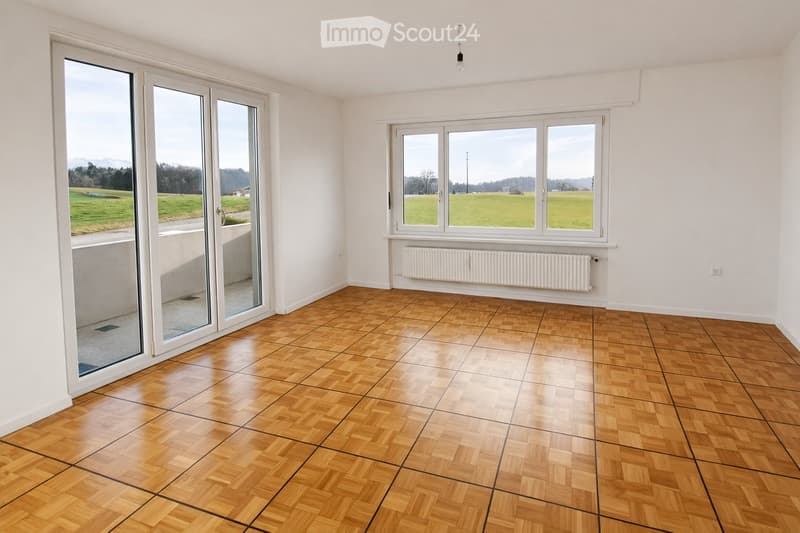 Empty room with wooden flooring, white walls, large windows, and a sliding glass door leading to a balcony.