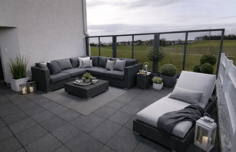 Balcony with grey wicker sofa, coffee table, sun lounger, potted plants, and lanterns.