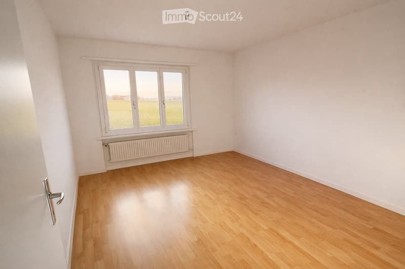 Empty room with light wooden flooring, white walls, and a large window with white frames. A radiator is visible below the window, and the room has a simple, clean design with no furniture or decorations.