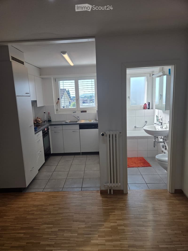 The image shows a modern, well-lit kitchen and bathroom area. The kitchen features white cabinets, a stainless steel sink, a dishwasher, and a refrigerator. The bathroom includes a bathtub, a sink, a toilet, and a mirror. Both areas have tiled floors, and