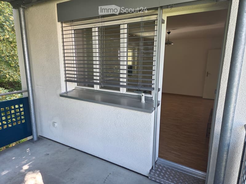 Balcony with a window featuring blinds, a small shelf, and a view of greenery outside. The interior shows a wooden floor and a doorway leading inside.