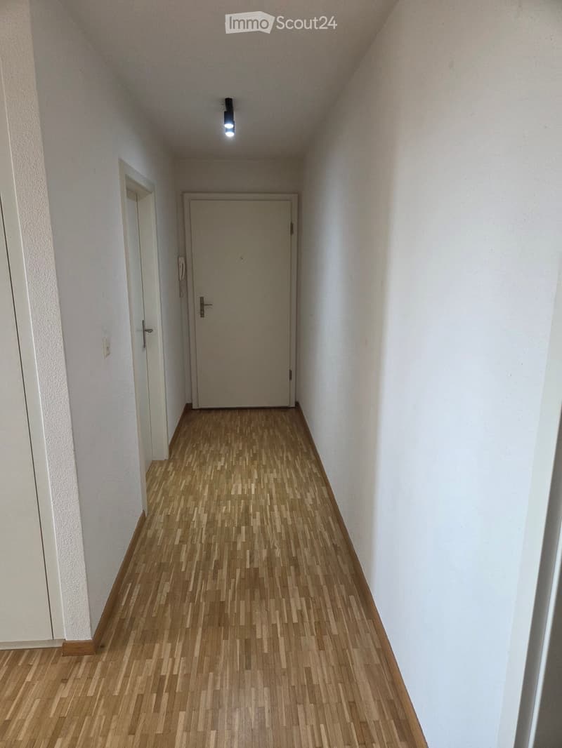 A narrow hallway with wooden flooring, white walls, and two doors, one on each side, illuminated by a single ceiling light.