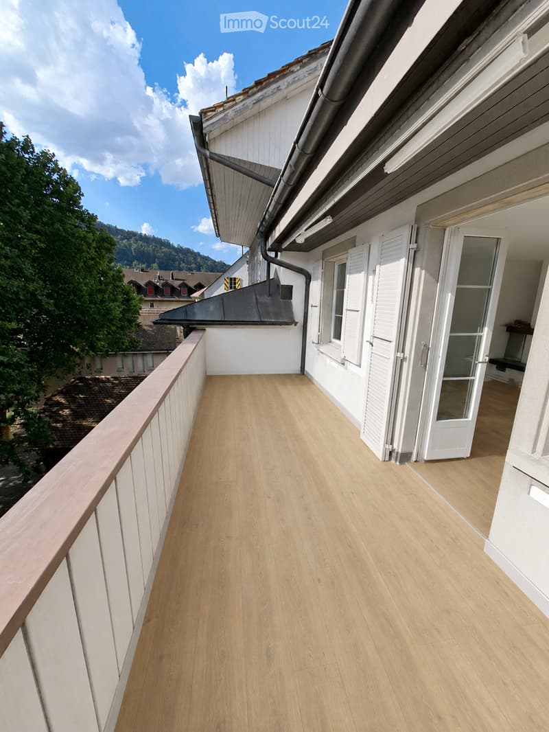 Balcony with wooden flooring, white walls, and white shutters. Features a wooden railing and access to the building interior.