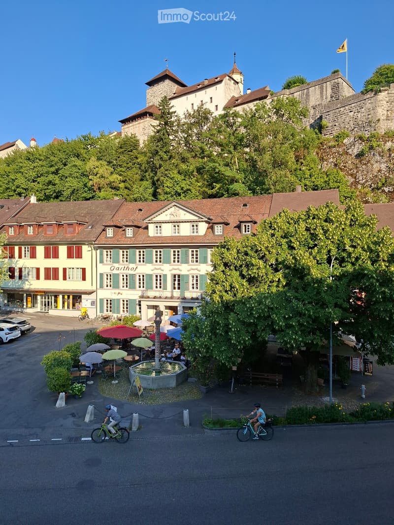 A town square with a fountain, outdoor seating, and historic buildings, surrounded by trees and a stone wall.