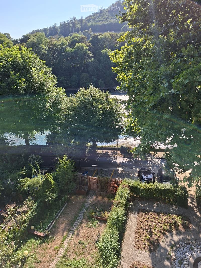 Aerial view of a lush garden area with trees, shrubs, a paved pathway, and a small wooden gate leading to a parking area with two cars.
