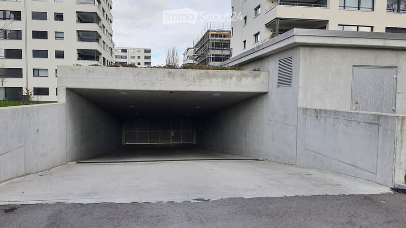 Modern concrete building entrance with a covered garage area, surrounded by other residential structures and greenery.