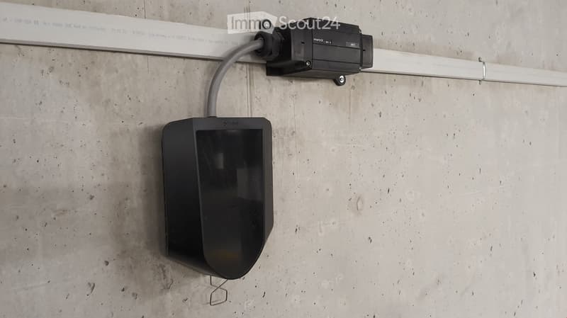 A modern electronic device mounted on a concrete wall, connected to a cable and a white rail system.