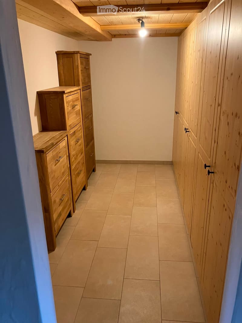 A narrow hallway with tiled flooring, wooden built-in cabinets on one side, and a wooden wardrobe on the other side. The ceiling has exposed wooden beams and a single ceiling light fixture.