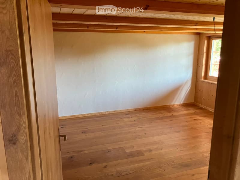 Empty room with wooden flooring and white walls, featuring exposed wooden beams on the ceiling and a small window letting in natural light.