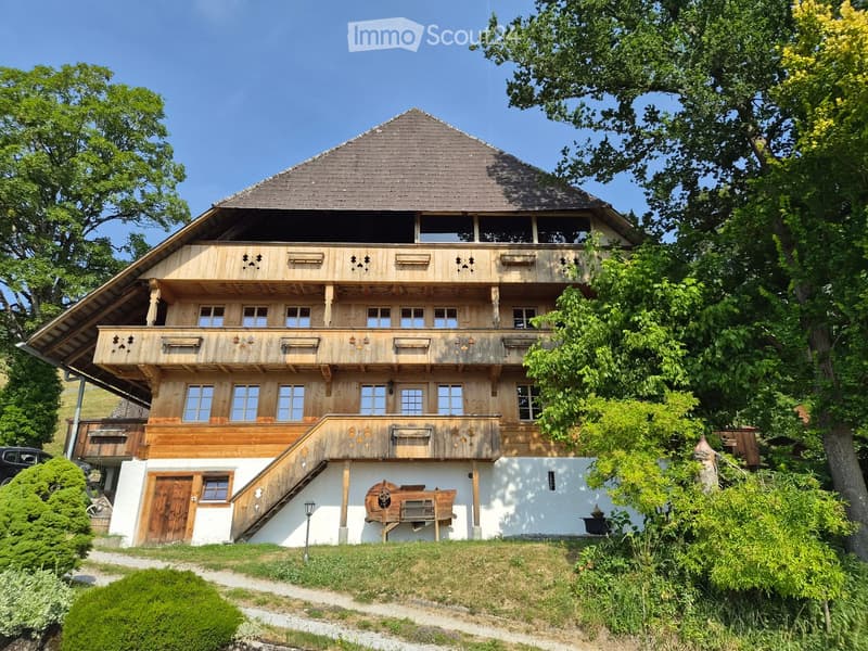 Traditional wooden house with a steep roof, multiple balconies, and decorative wooden trim. The house has a raised ground floor and is surrounded by greenery and trees.