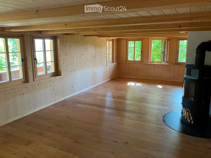 Empty room with wooden walls, wooden floor, large windows, and a wood-burning stove with logs nearby.