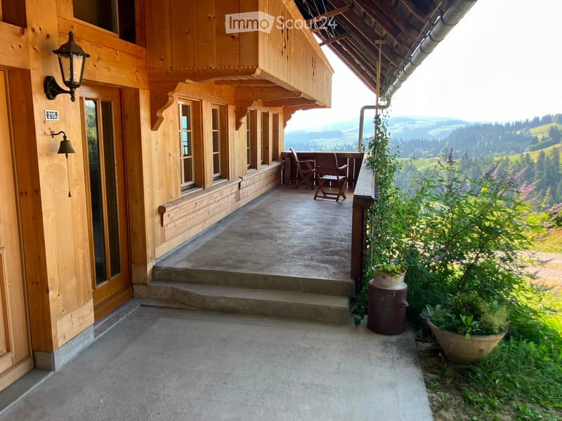 Wooden balcony with chairs and a small table, overlooking a scenic valley with hills and trees.