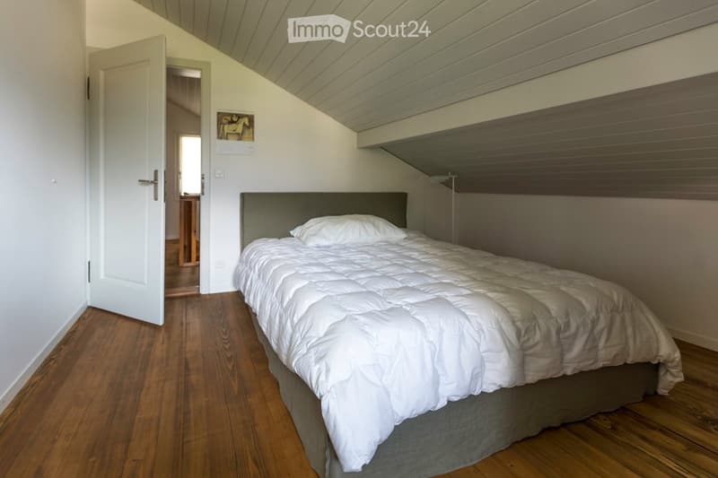 Cozy bedroom with wooden flooring, a neatly made bed with white bedding and pillows, and a dark grey headboard. An open door leads to another room.