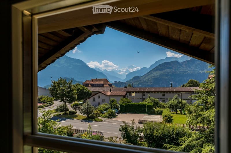 View from a balcony showing a town with buildings, trees, and a road, with snow-capped mountains in the background.