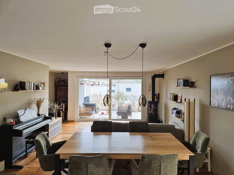 Bright living and dining area with wooden flooring, large windows, a sofa, dining table with chairs, piano, and decorative shelves.