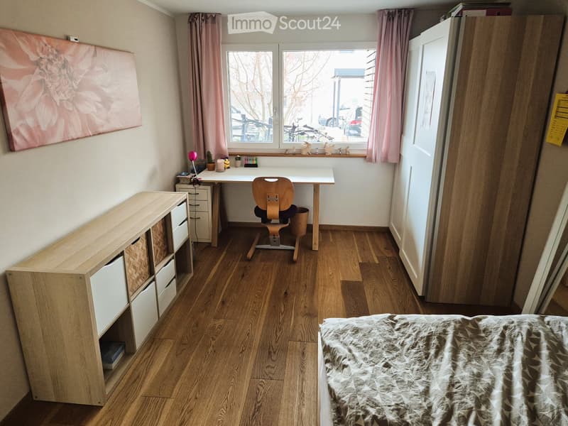 Cozy bedroom with wooden flooring, a bed with a patterned blanket, a wooden desk with a chair, a storage unit, and a cabinet with drawers.