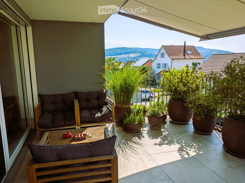 Balcony with a sofa, wooden table, chair, and several potted plants, offering a scenic view of the surrounding area.